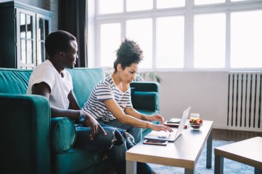 Siyahi adamın odaklanmış kız arkadaşına dizüstü bilgisayar kullanarak bakarken evdeki pencereye karşı rahat bir koltukta oturması.