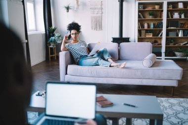 Gözlüklü, akıllı telefonlu, kağıt taşıyan ve modern apartmandaki koltukta çıplak ayakla oturan Afrikalı Amerikalı bir kadın. 