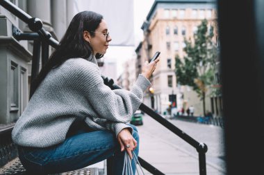 Gri kazaklı ve kot pantolonlu bir kadın sokak merdivenlerinde otururken New York 'un caddesinde akıllı telefon kullanıyor. 