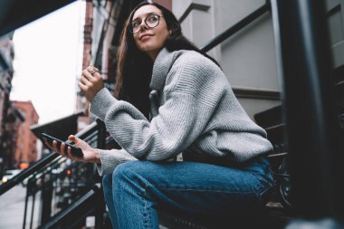 Gri kazaklı, mavi kot pantolonlu, akıllı telefonları karıştıran ve New York sokaklarında gülümseyen düşünceli genç bir kadının yan görüntüsü. 