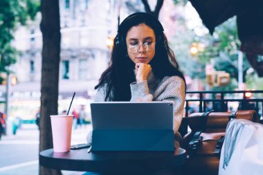 Genç, düşünceli, ince kadın, gözlüklü ve kulaklıklı, elleri çenesinin altında, kafein bardağının dışında karton bardakla masada gezinen.