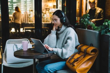 Gri kazaklı, turuncu çantalı, gözlüklü, kulaklık takan ve New York Kafe 'de dijital tablet üzerinde çalışan serbest bir genç kadın. 