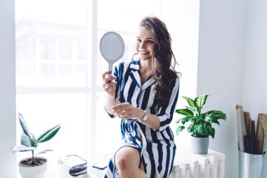 Moda kıyafetleri içinde çekici bir kadın pencere pervazında oturuyor ve evde ayna ve kozmetik ürünleriyle tuvalet yapıyor.