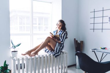 Düşünceli kıvırcık saçlı, çizgili elbiseli, kitap okuyan ve güneşli bir günde şık bir ev ofisinde pencere eşiğinde oturan esmer.