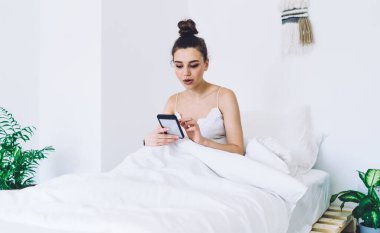 Odaklanmış bir kadın, beyaz battaniye ve yastık ile rahat yumuşak yatak üzerinde uyanır ve ışık odasında minimalist iç mekan ile akıllı telefon sörfü yapar.