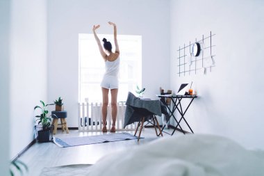 Çıplak ayakla esinlenmiş pijamalı bir kadın ellerini kaldırıp modern odadaki beyaz duvarlara bakıyor.