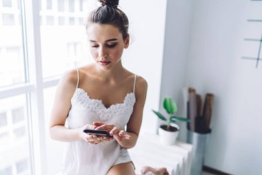 Zarif ipek gecelikler giymiş sevimli genç bir bayan. Açık iş dantelli, akıllı telefonuyla sohbet ederken açık beyaz odada pencere eşiğinde oturuyor.