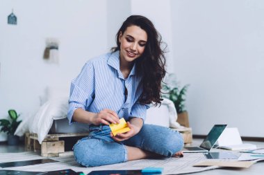 Çekici, uzun saçlı, sarı etiketli bir kadın. Siyah resimlere yapıştırıyor. Dizüstü bilgisayarlı, aydınlık odada oturuyor.