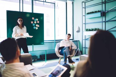 Modern konferans odasında şirket stratejileri hakkında konferans verirken gündelik giysiler içindeki kendine güvenen genç çekici yönetici yazı tahtasının yanında duruyor.