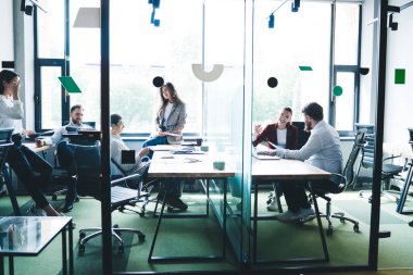 Masada dizüstü bilgisayarlı ve çalışma kağıtlı neşeli ve konsantre iş arkadaşları sohbet ediyor ve cam ofis odasında birlikte çalışıyorlar.