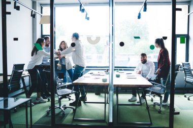 Elleri üst üste dizilmiş bir grup mutlu iş arkadaşı ofiste cam duvarla durup gülümserken başarıya seviniyorlar.
