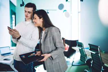 Modern çalışma odasında masanın yanında duran ve kağıt üzerinde diyagramları analiz ederken telefon izleyen genç pozitif şirket çalışanları