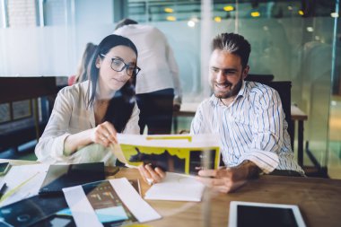 Ofis arkadaşları, modern çalışma alanındaki ofis malzemeleriyle cam arkasında otururken işle ilgili bilgilerle kağıt sayfalarını tartışıyorlar.