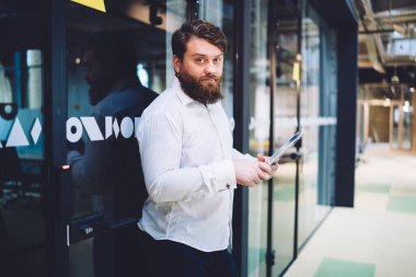 Ciddi sakallı bir yönetici, modern ofis binasında cam kapıya yaslanırken kameraya bakıyor ve tablet üzerinde çalışıyor.