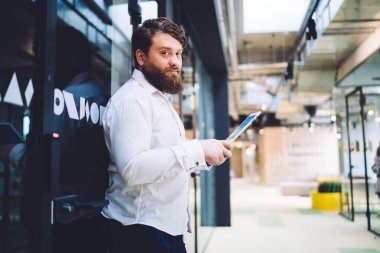 Modern ofis binasında cam kapıya yaslanırken kameraya bakan ve tablet üzerinde çalışan ciddi sakallı erkek yöneticinin yan görüntüsü bulanık arka planda.