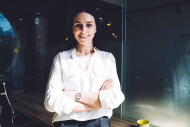 Parlak odada kollarını kavuşturup kameraya bakarken gülümseyen resmi giyinmiş neşeli ve mutlu bir bayan.