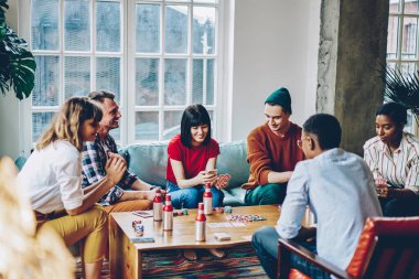 Kanepede oturup arkadaşlarıyla poker oynarken, akıllı telefondan gülümseyen ve kombinasyonu kontrol eden gündelik kıyafetli mutlu genç bir kadın.