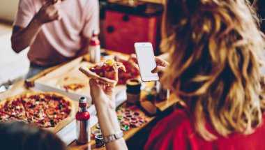 Yukarıdan gelen iştah açıcı pizza dilimi cep telefonuyla fotoğraf çeken bir grup çok ırklı insanla aynı masada yemek yiyor.
