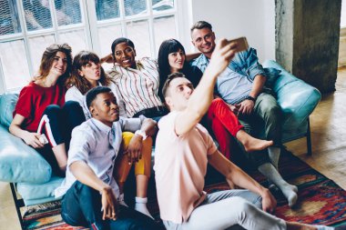 Yukardan gelen mutlu milenyum çeşitliliği öğrencileri günlük kıyafetleri içinde rahat modern apartman dairesinde buluşurken akıllı telefonlarıyla selfie çekiyorlar.