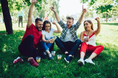 Parkta güneşli bir günde çimlerde otururken moda kıyafetleri içinde video izleyip çığlıklar atan bir grup genç. 