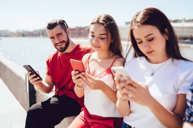 Modern akıllı telefonlu yakışıklı sakallı adam güneşli bir günde sette bayan arkadaşlarıyla takılırken gülümsüyor ve kameraya bakıyor.