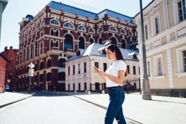 Beyaz gömlekli, kot pantolonlu, güneşli bir yaz gününde caddeden karşıya geçerken ekranda akıllı telefonla etkileşen muhteşem sakin esmerin alçak açılı görüntüsü.