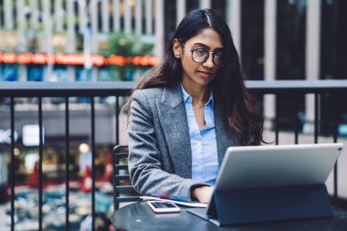 Genç odaklı gözlüklü, belgelerle çalışan ve dışarıdaki terasta kentsel arka plan üzerine masaya oturmuş tablet izleyen etnik bir kadın.