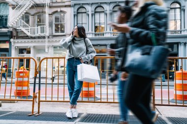 Gri elbiseli, kot pantolonlu, genç bir kadın sokakta çitlere yaslanıyor, alışveriş çantaları alıyor ve New York 'ta cep telefonundan arıyor.