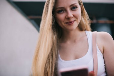 Düşük açılı, beyaz kolsuz tişörtlü, mutlu, genç bir kadın şehirde cep telefonuyla kameraya bakıyor.