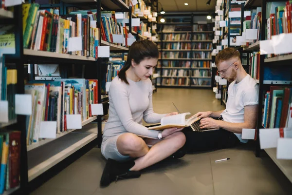 专注的女生坐在图书馆的地板上看书 红头发大胡子的男生靠在书架上用笔记本电脑 — 图库照片