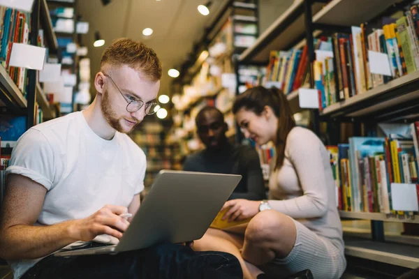 Focused ginger man watching laptop enthusiastically while studying with intelligent multiethnic students among high shelves with books on library floor