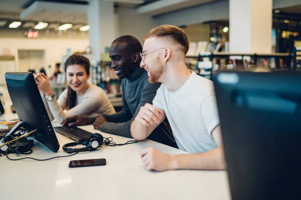 Side view of multiracial laughing active students using computer in library and talking joyfully while preparing for college exams together