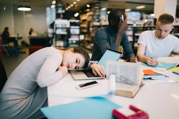 Sleepy woman dressed in casual clothes napping on laptop on table with notepads near executives discussing project in office building
