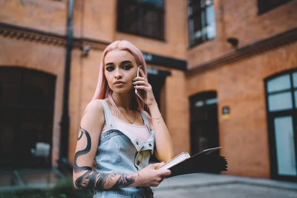 Stylish young lady with snake tattoo and long pink hair dressed in jeans dress speaking on mobile phone holding book in hand
