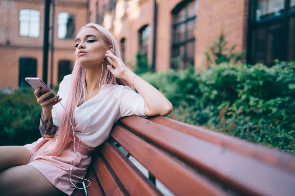 Sensual young lady in white shirt and pink shorts with long pink hair listening to music on smartphone with earphones sitting on wooden bench in street