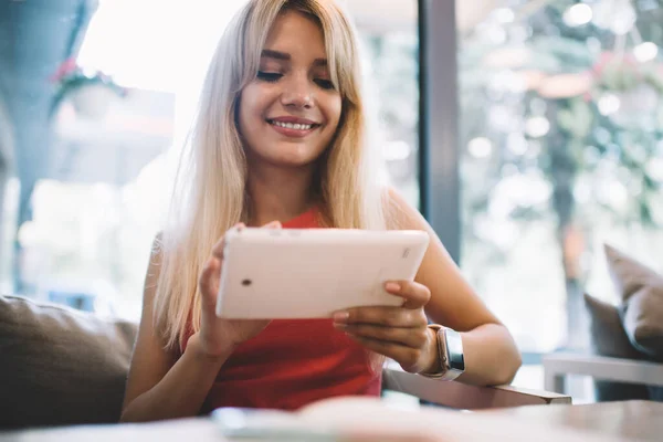 在舒适的咖啡馆里 年轻的金发女子坐在靠窗的沙发上 开心地微笑着 用现代平板电脑 — 图库照片