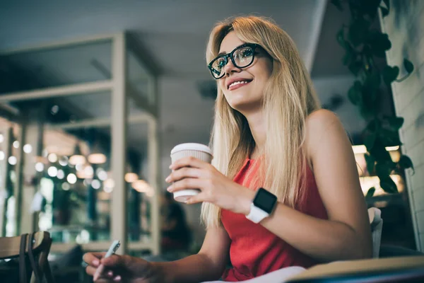 From below cheerful youthful woman with light hair wearing glasses and drinking takeaway beverage while making notes and sitting in cafe