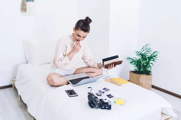 Worried thoughtful adult woman in casual clothes biting nails focusing on screen while sitting crossed legs with opened notepad in hand and using laptop on bed in minimalistic white bedroom