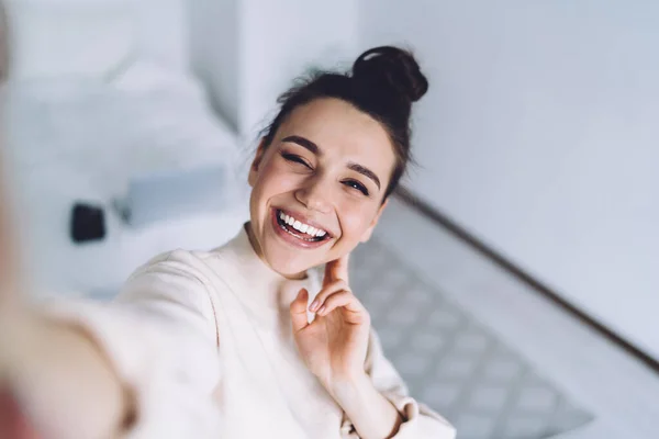 Winking joyful lady with golden smile with hand beside face laughing and taking selfie on mobile phone at light bedroom on blurred background