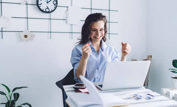 Pretty adult woman in blue shirt and glasses with raised arms concentrating on laptop screen while sitting at desk against white wall with black decor and clock