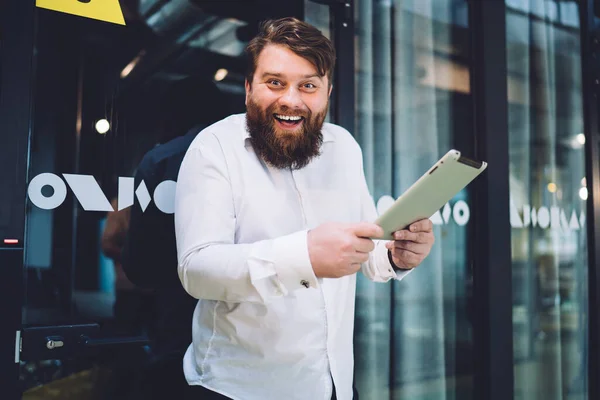 Joyful bearded male office employee holding tablet with good news on it while looking at camera leaning against glass door in hallway of modern office