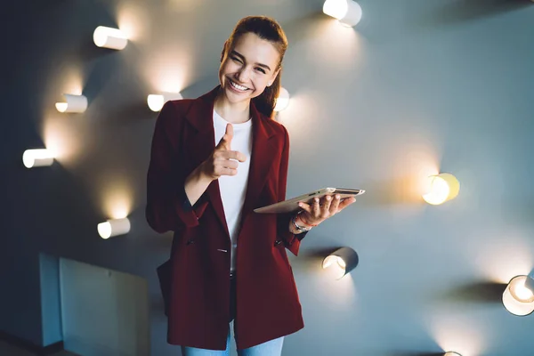 Friendly female employer in stylish red coat with tablet winking and pointing with index finger at camera while standing near wall with decorative lighting