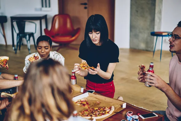 Young multiracial company of friends in casual clothes eating pizza and drinking beer while playing table games during party at apartment