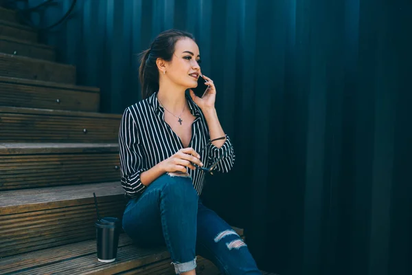 青い縞模様の壁に対して木製の階段に座っている間に電話を作るためにサングラスや飲料とカジュアルな服の中で高価な自信の女性の低角度 — ストック写真