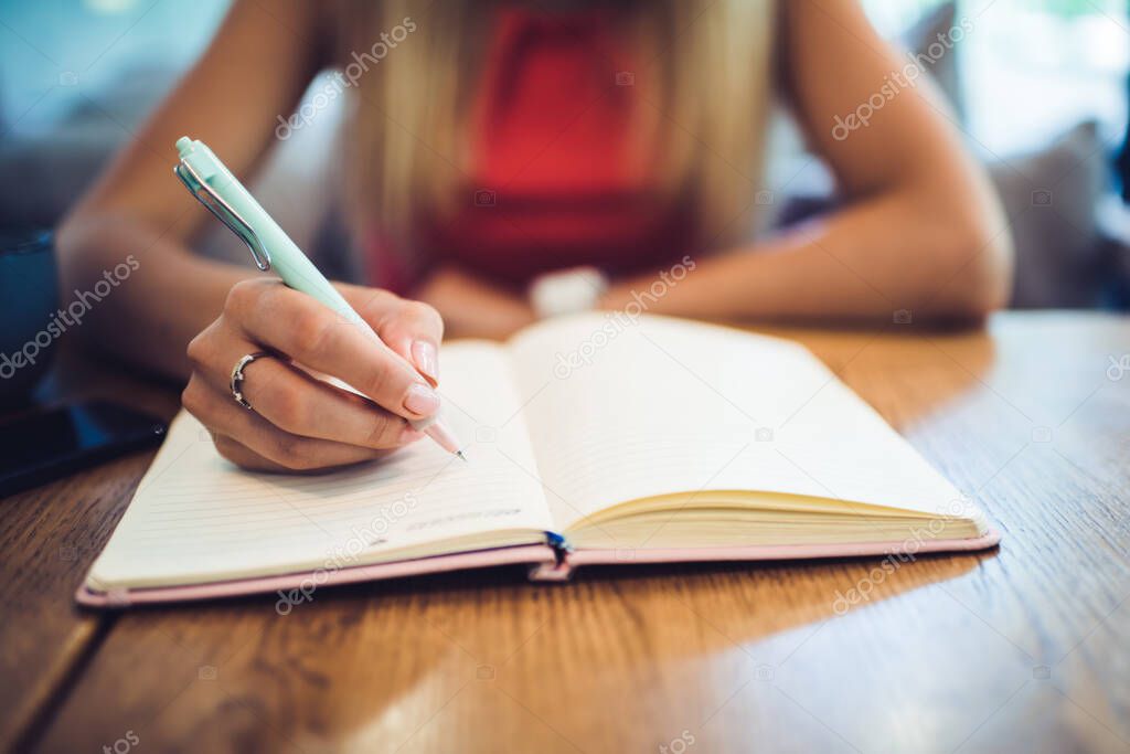 Unrecognizable female using pen to make notes in open notepad while sitting at table and resting in modern cozy cafe