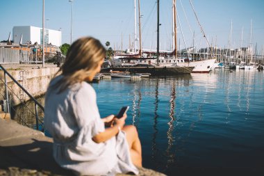 Bin yıllık hipster kızının arama bilgileri ve internet sitesi için akıllı telefon aygıtı kullanarak limanda dinlenirken görüntüsü bulanık yüzlü deniz kenarında oturan bir kadın.