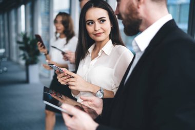 Güzel, genç bir bayan, mesai arasında iş arkadaşlarının yanında dijital cihazlara bakarken gülümsüyor ve erkek iş arkadaşına bakıyor.