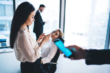 Beyaz masaya yaslanmış siyah saçlı ve ofis çalışanlarının yanındaki akıllı telefondan pencerelere doğru mesaj atan iş adamının yan görüntüsü 