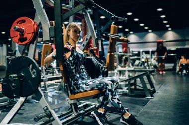 Sağlıklı, güçlü, kararlı, gri kamuflaj elbiseli sporcu kadın modern spor salonunda göğüs makinesiyle oturup ağırlık kaldırıyor.