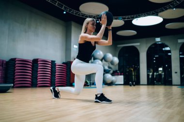 Düşük açılı, siyah üst ve beyaz taytlı güzel kadın spor salonunda tıp topuyla atlıyor ve başka yöne bakıyor.
